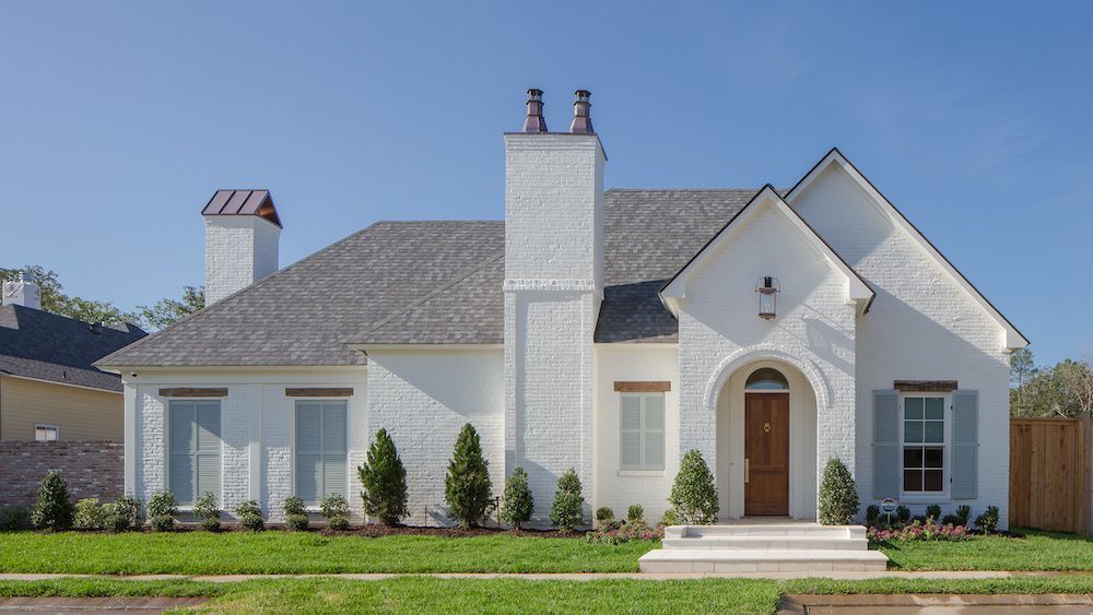 A white, one-story house with a brick exterior, prominent chimney, gray roof, and a central arched wooden front door.