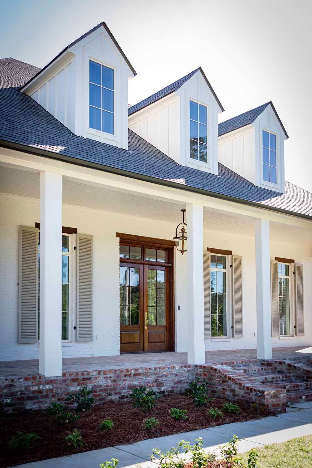 A white, single-story house featuring a brick porch, large wooden front doors, and three prominent roof dormers.