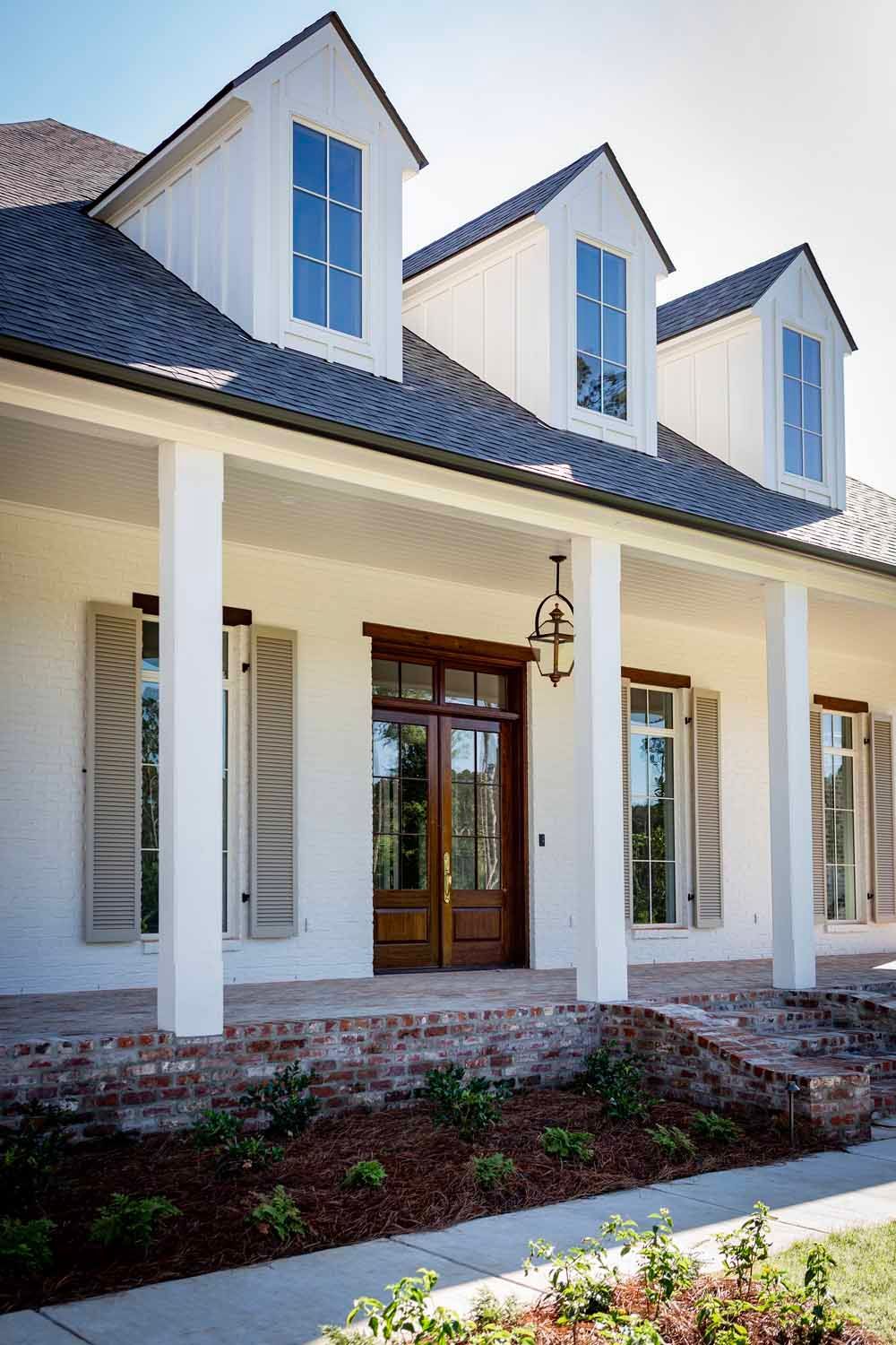 White house facade with three dormers, a covered porch, brick skirting, and a dark wooden front door.
