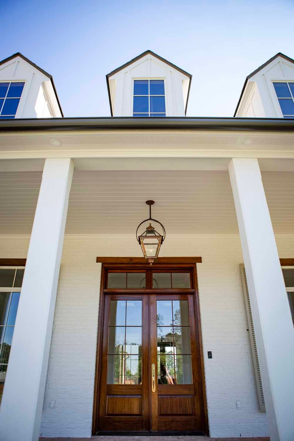 A low-angle view of a white brick home entrance with tall columns, a wooden double door, a hanging lantern, and dormers.