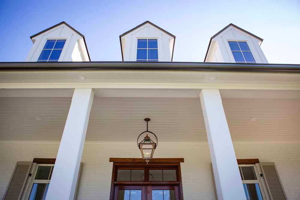 Low-angle view of a white building with three dormer windows, a covered porch with large pillars, and a hanging lantern.