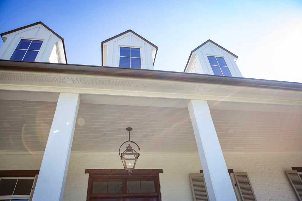 A low-angle view of a white building exterior with a porch, columns, hanging lantern, and three white attic dormer windows.