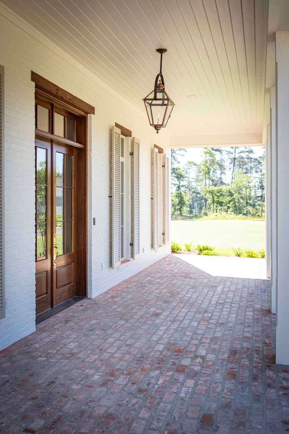 A covered brick porch with a wooden door, cream shutters, and a hanging lantern, leading to an open outdoor area.