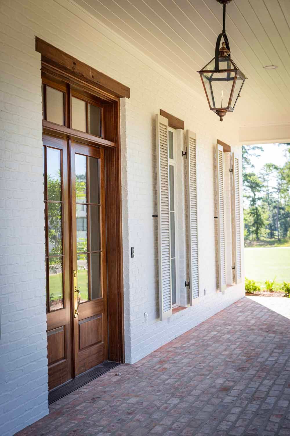 A white brick porch with tall wooden doors and shuttered windows, lit by a hanging lantern over a red brick floor.