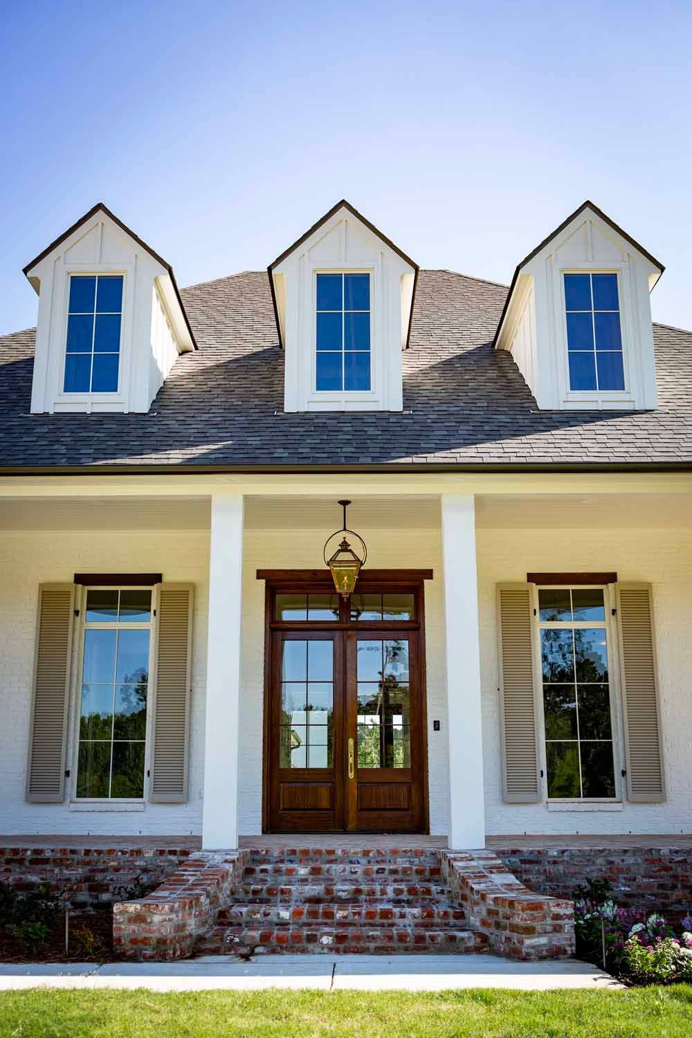A white house with a brown front door, symmetrical windows with shutters, three gabled dormers, and brick stairs.