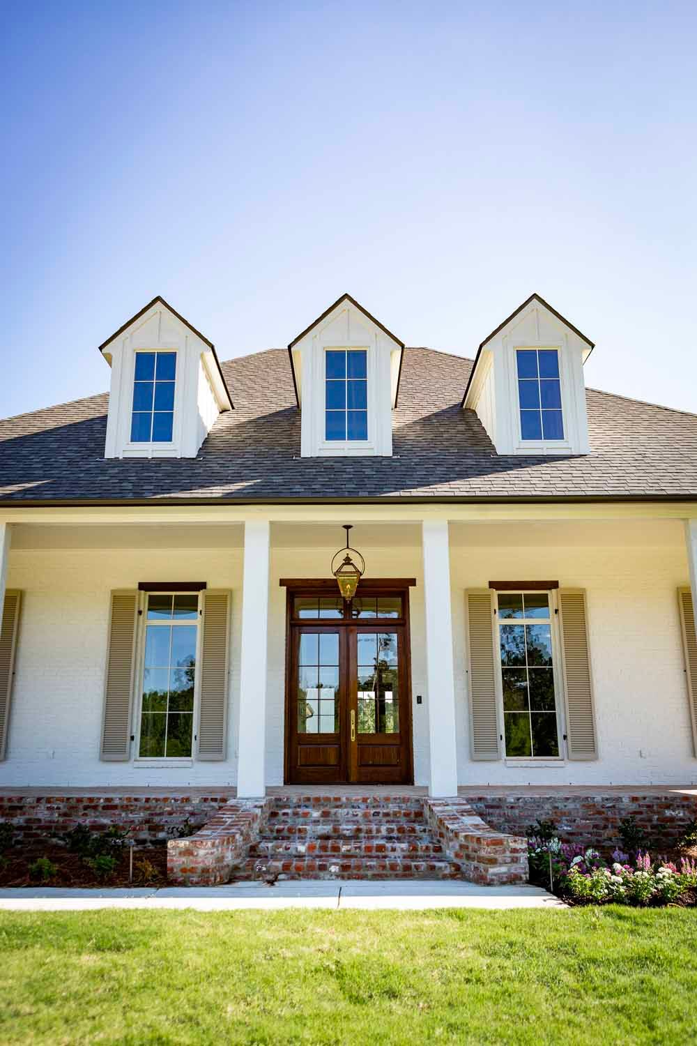 A single-story white house with a gray roof, three dormer windows, a brick base, and a wooden front door under a porch.