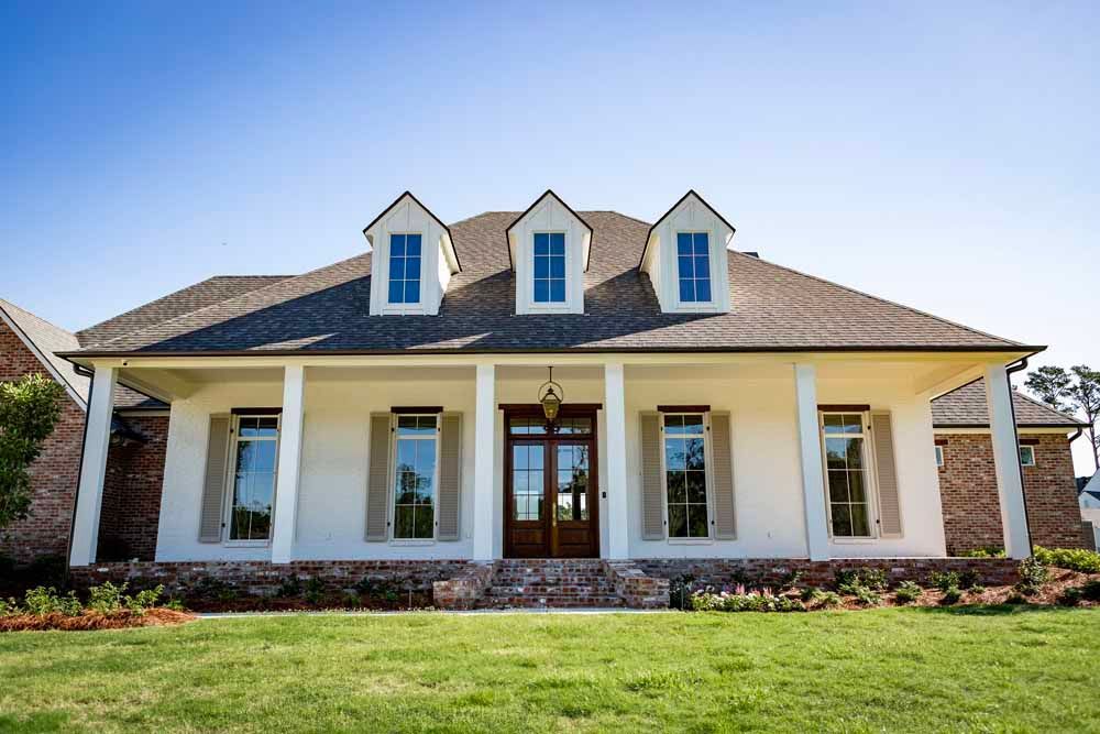A white, single-story house with a brick base, dark roof, three dormer windows, and a wide front porch under a clear sky.