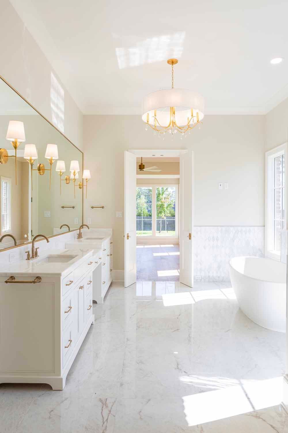 A bright, white bathroom featuring marble floors, a double vanity, a freestanding tub, and a decorative gold chandelier.