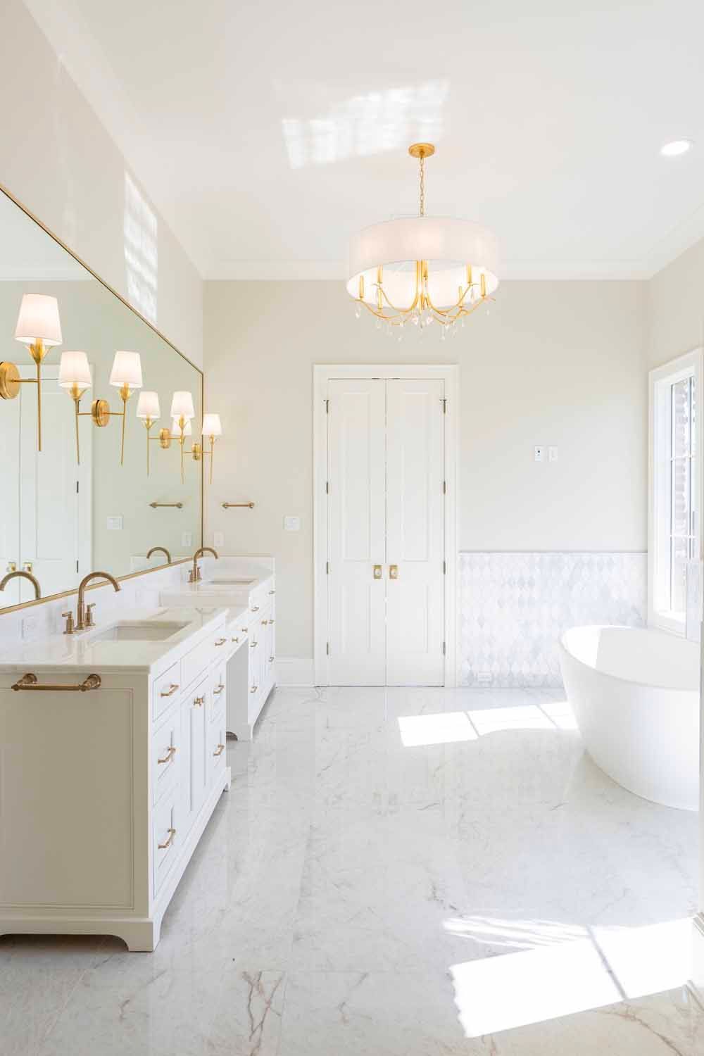 Bright, modern bathroom featuring a white vanity with gold hardware, marble flooring, a soaking tub, and gold lighting.
