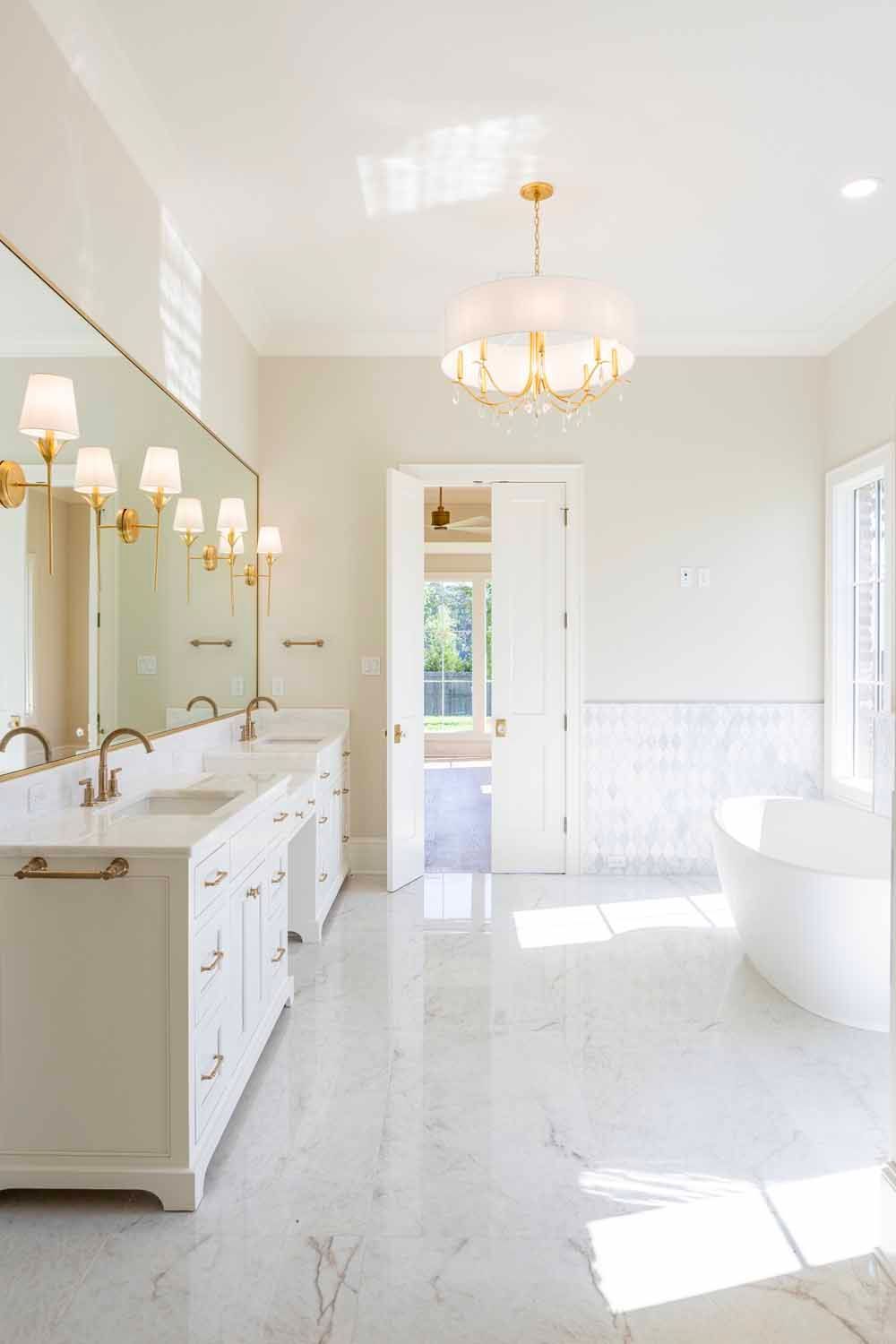 A bright, modern white bathroom with marble floors, a double vanity, a freestanding tub, and a gold chandelier.