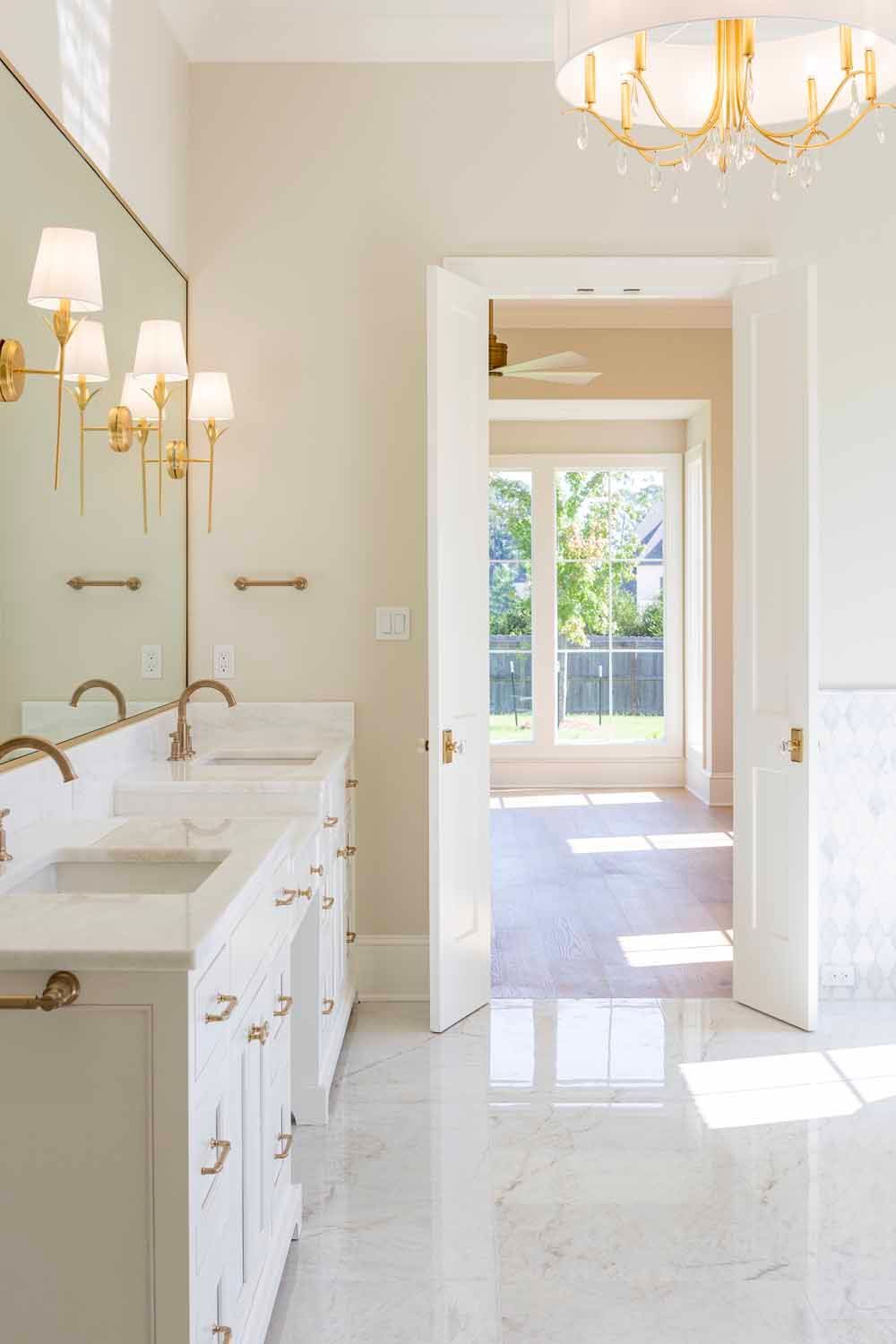 A bright bathroom with white vanity cabinets, marble floors, gold fixtures, and double doors opening to a sunlit room.