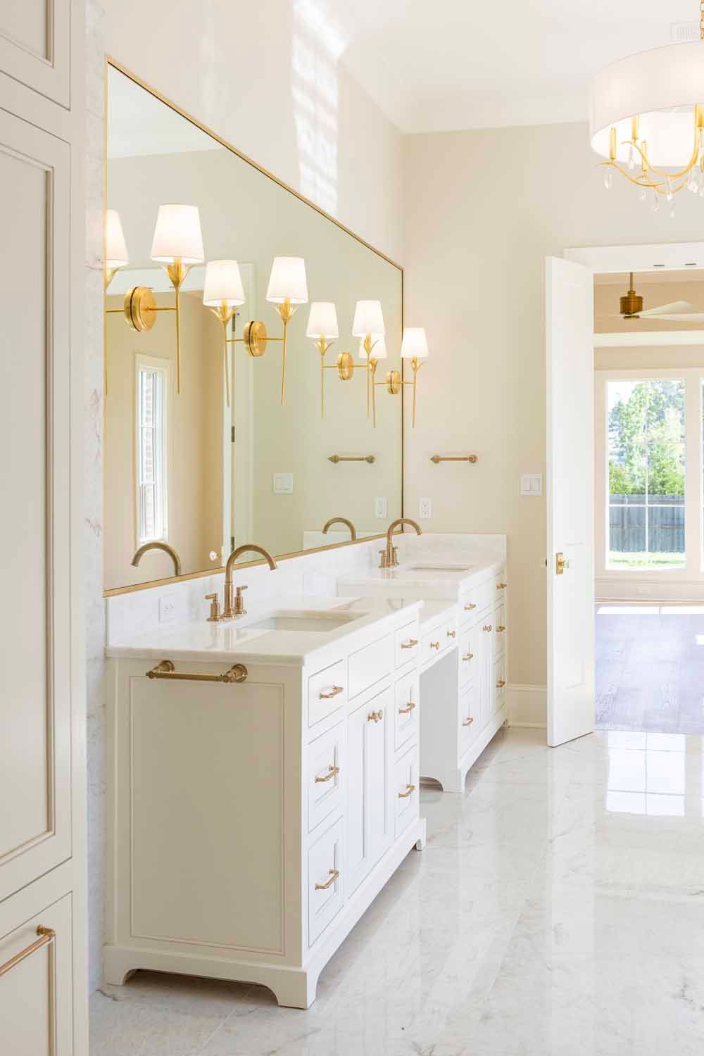 A bright, white bathroom vanity with gold hardware, double sinks, and a large mirror with wall-mounted lamps.