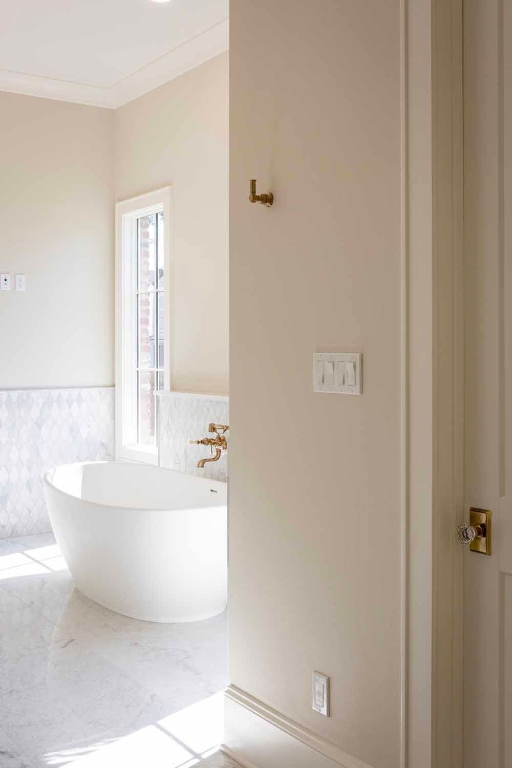 A bright, modern bathroom featuring a white soaking tub, marble flooring, and light-colored walls with gold fixtures.