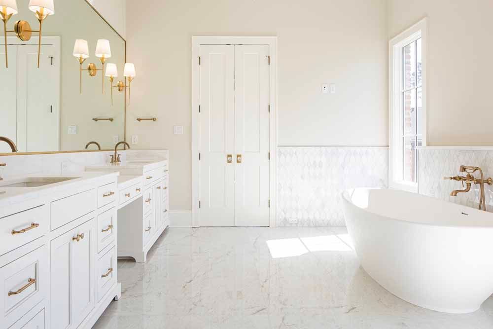A bright, white bathroom with a freestanding tub, marble floors, a double vanity, and tall double doors.