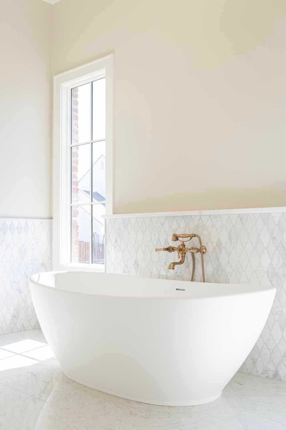 A white freestanding bathtub with brass hardware against a white tiled wall in a sunlit bathroom.