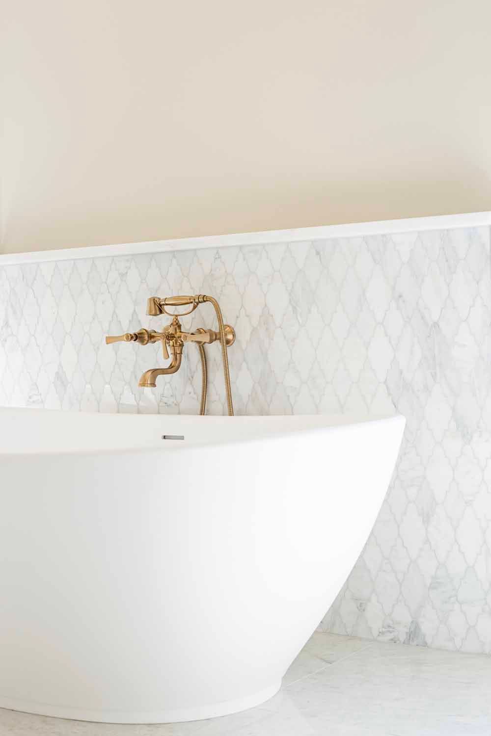 A white freestanding bathtub with a gold vintage faucet against a white marble diamond-patterned tile wall.