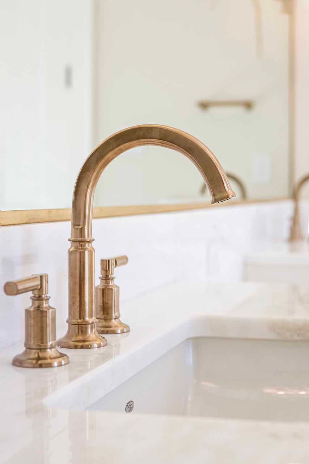 A gold-toned, modern bridge bathroom faucet with two handles, installed on a white marble countertop.