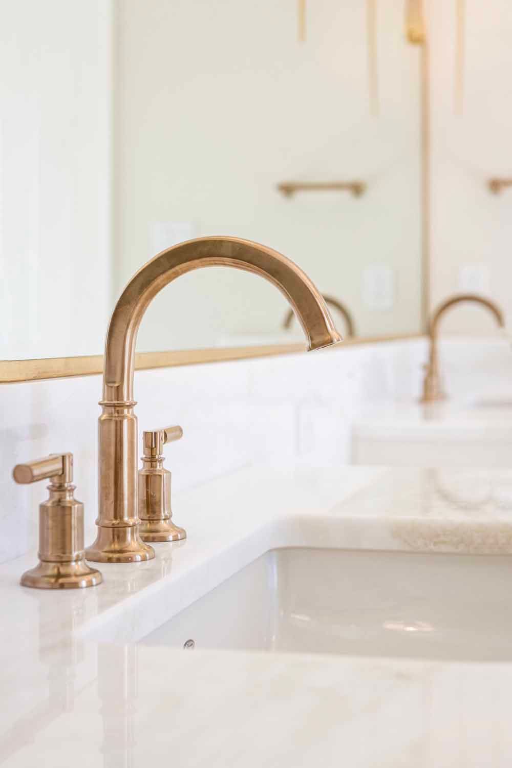 A close-up of a brushed gold bathroom faucet and handles on a white marble vanity, with a mirror reflecting the room.