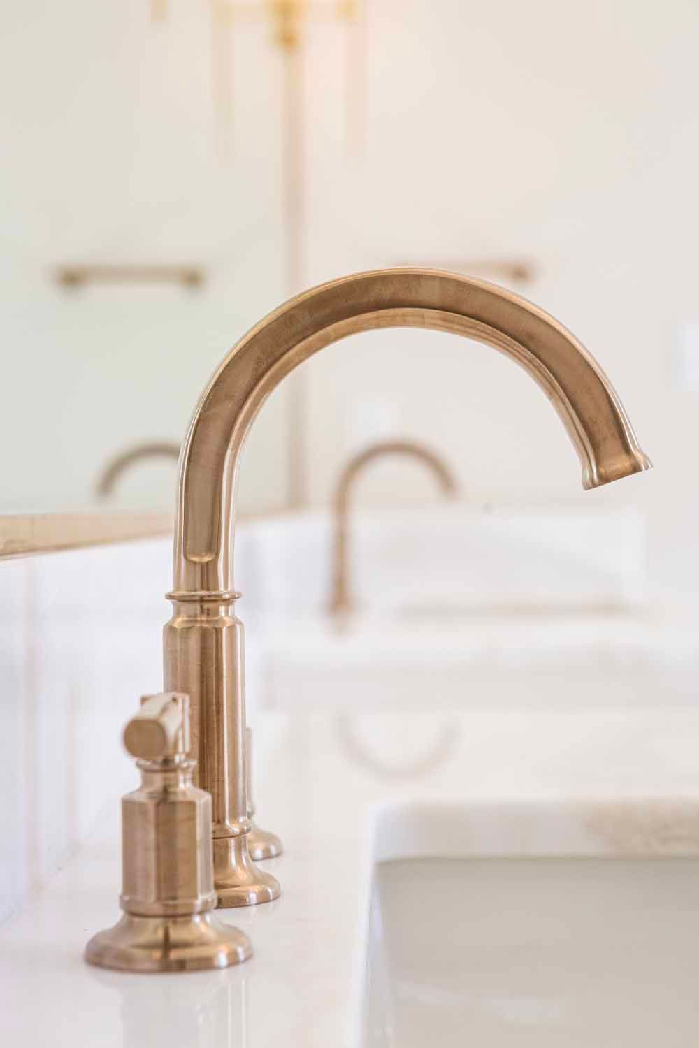 A sleek, gold-toned bathroom faucet with a side handle stands on a white countertop in a bright room.
