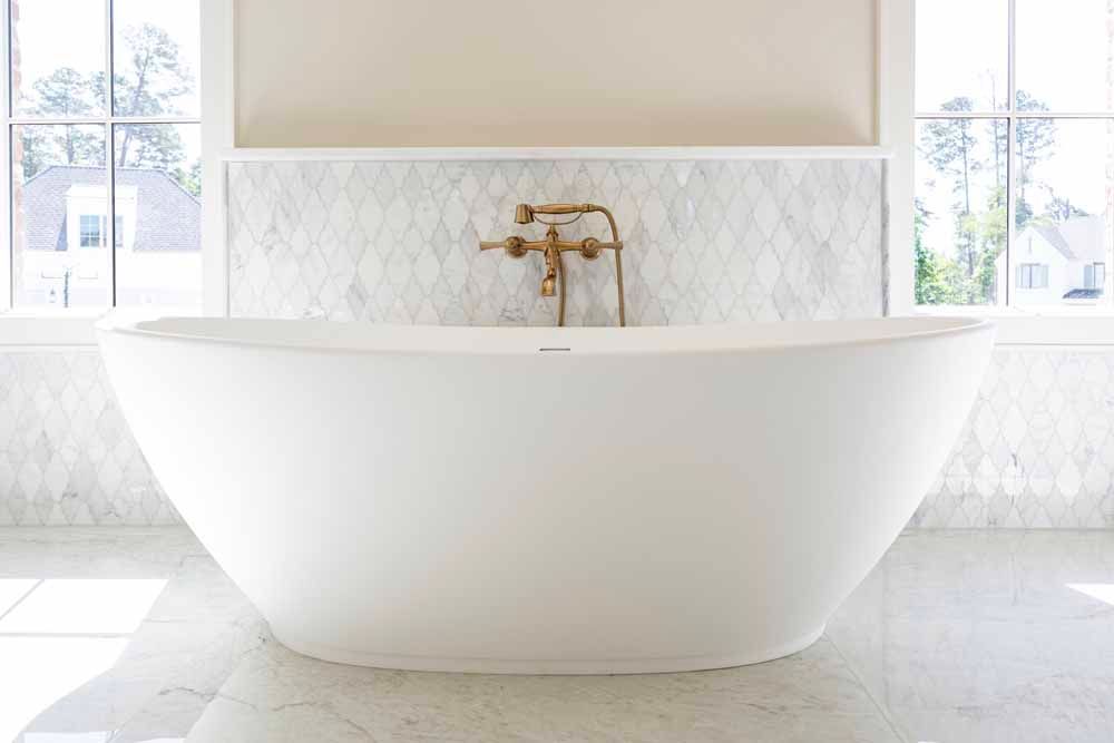 A white, freestanding bathtub centered in a bright bathroom with gold hardware and white diamond-patterned tile walls.