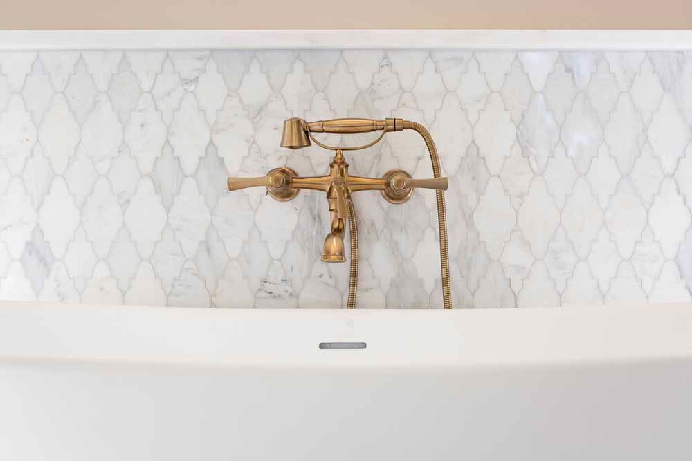 A brass bathtub faucet with a hand shower mounted against a white marble wall with a diamond-pattern tile backsplash.