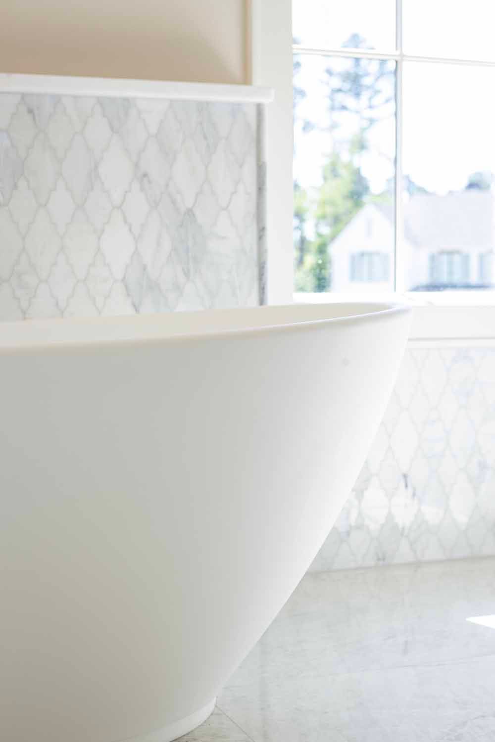 A sleek, white freestanding bathtub sits in a brightly lit bathroom with marble diamond-patterned wall tiles.