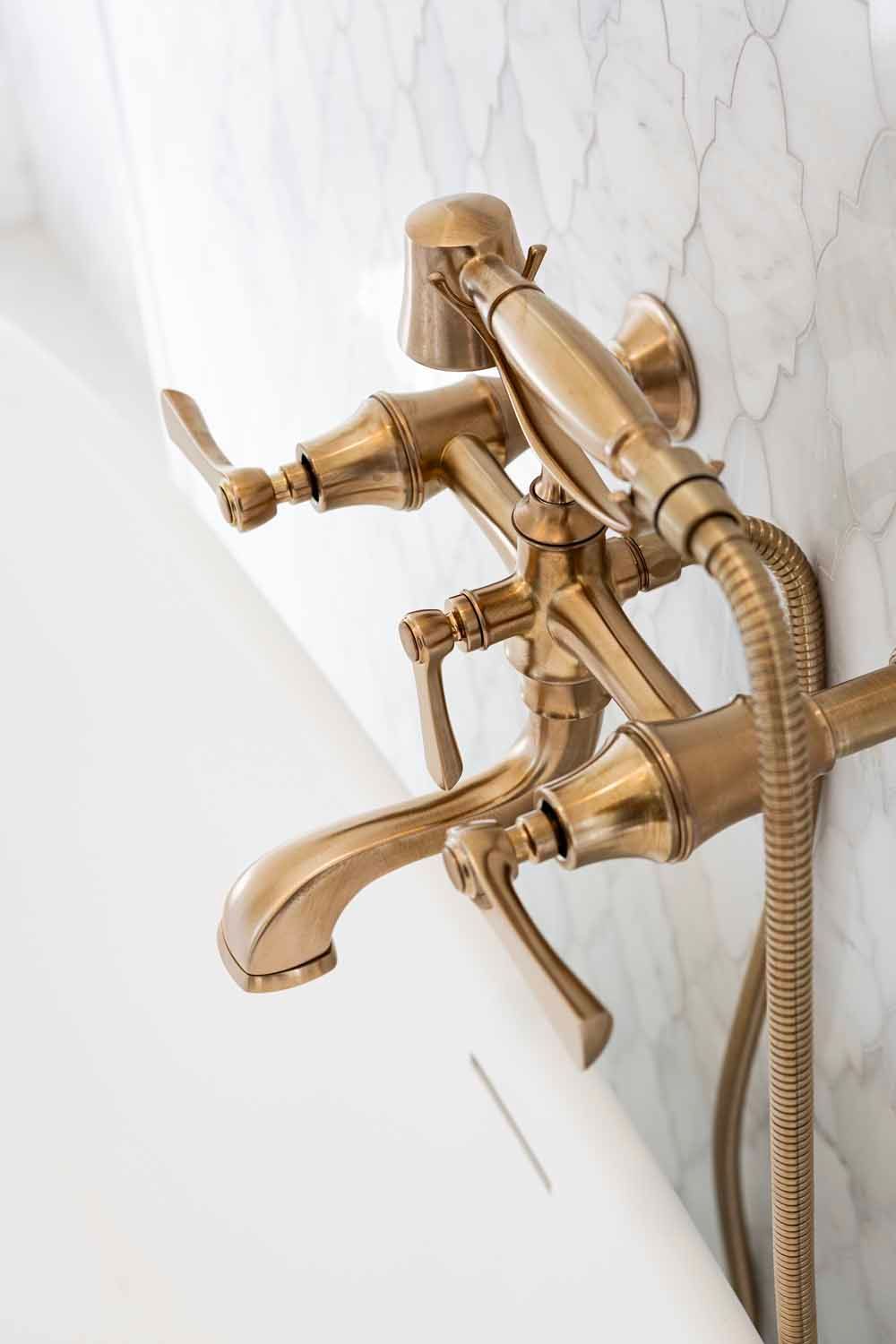 A brass wall-mounted bathtub faucet with a handheld shower attachment, set against a white marble tile wall.