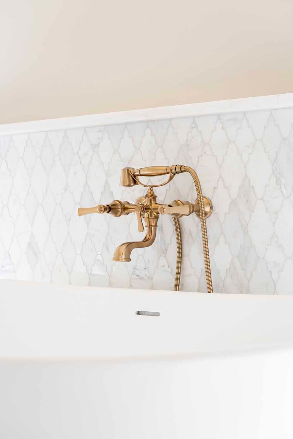 A brass vintage-style bathtub faucet and handheld shower sprayer mounted against a white diamond-patterned marble wall.