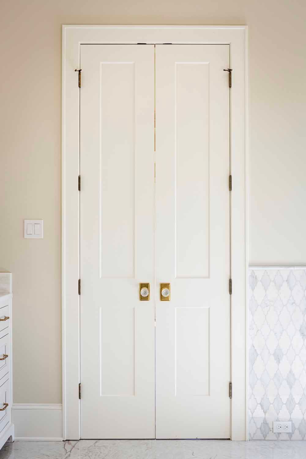A set of double white interior doors with brass knobs and hinges, set within a neutral-colored room.