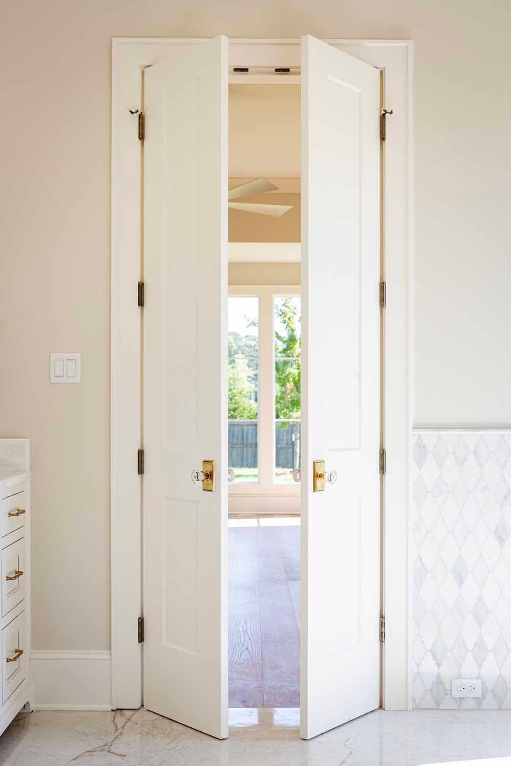 A pair of white double doors with gold knobs, slightly ajar, opening into a sunlit room with a large window.