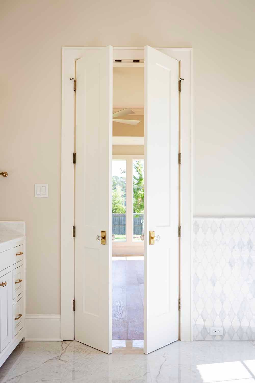 A set of white double doors with gold knobs, slightly ajar, standing in a brightly lit room with a tiled floor.
