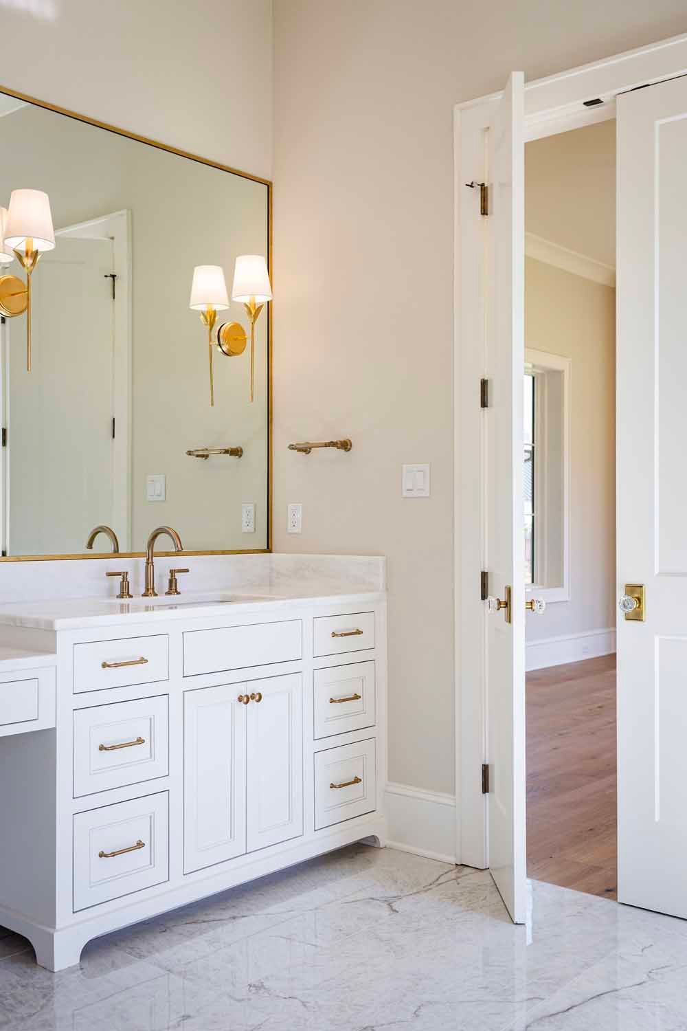 A bright, modern bathroom featuring a white double vanity, gold-rimmed mirror, wall sconces, and a white door left ajar.