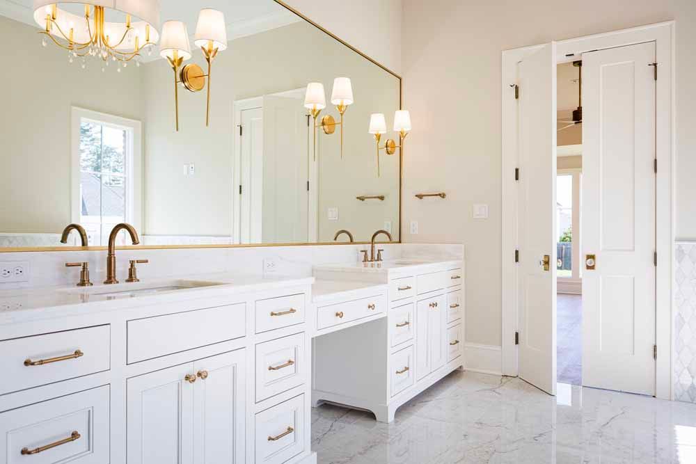A bright, white bathroom vanity with marble floors, gold fixtures, and a large mirror with gold wall sconces.