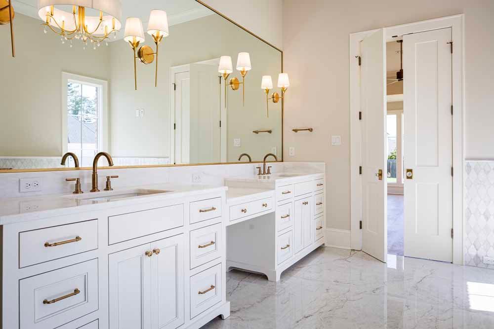 A bright, modern bathroom with white double vanities, marble countertops, brass fixtures, a large mirror, and open doors.