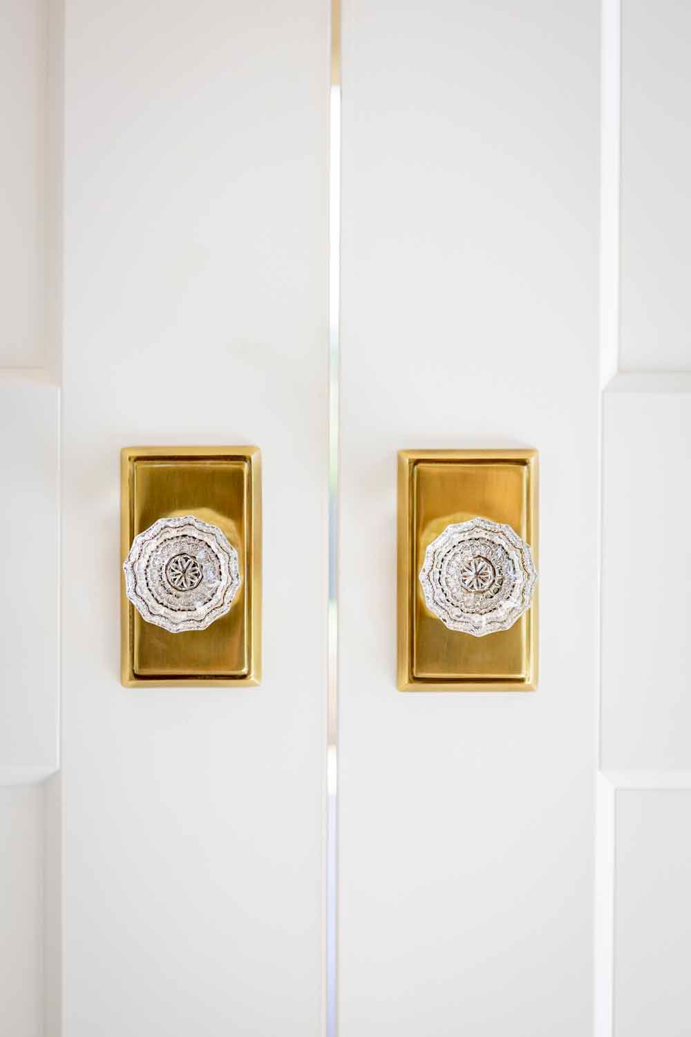 Two white doors side-by-side, each featuring a vintage gold rectangular plate with a round, clear crystal knob.