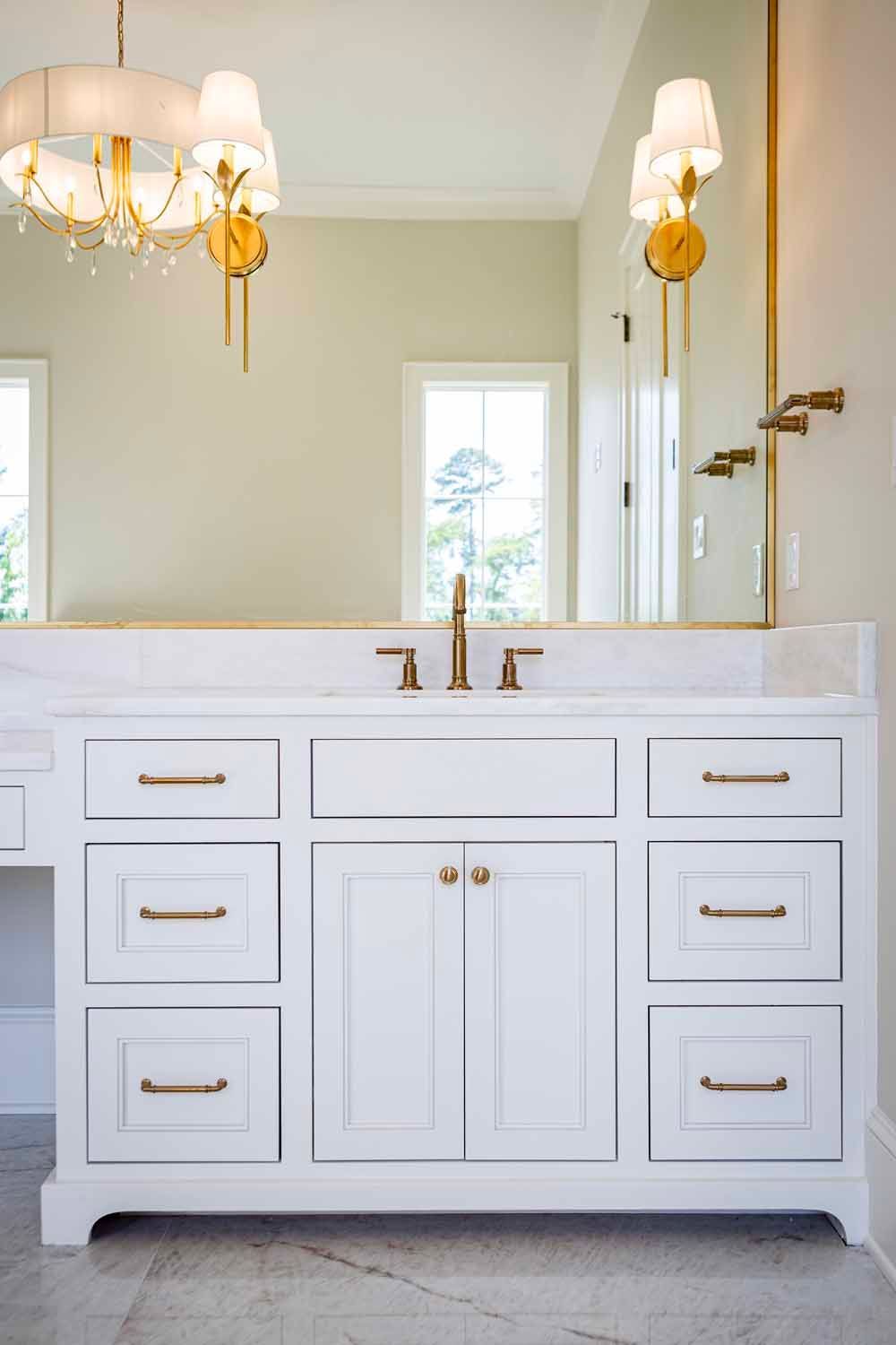 White bathroom vanity with gold hardware, a marble countertop, and a large mirror with gold-tone sconces and a chandelier.