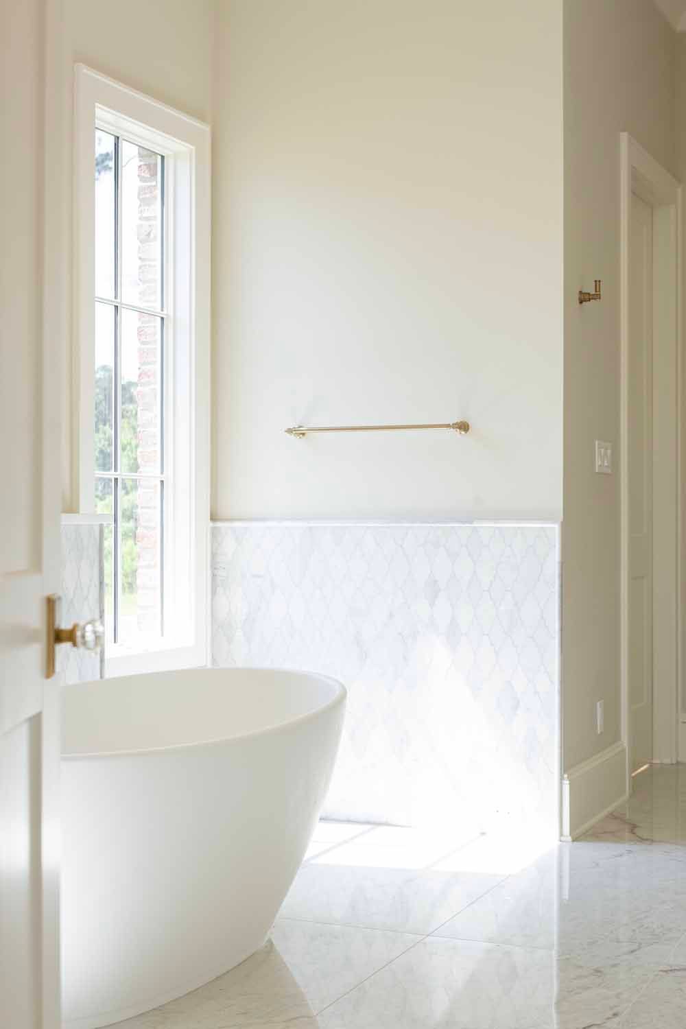 A white freestanding bathtub sits in a sunlit bathroom with white marble floors and wall tile.