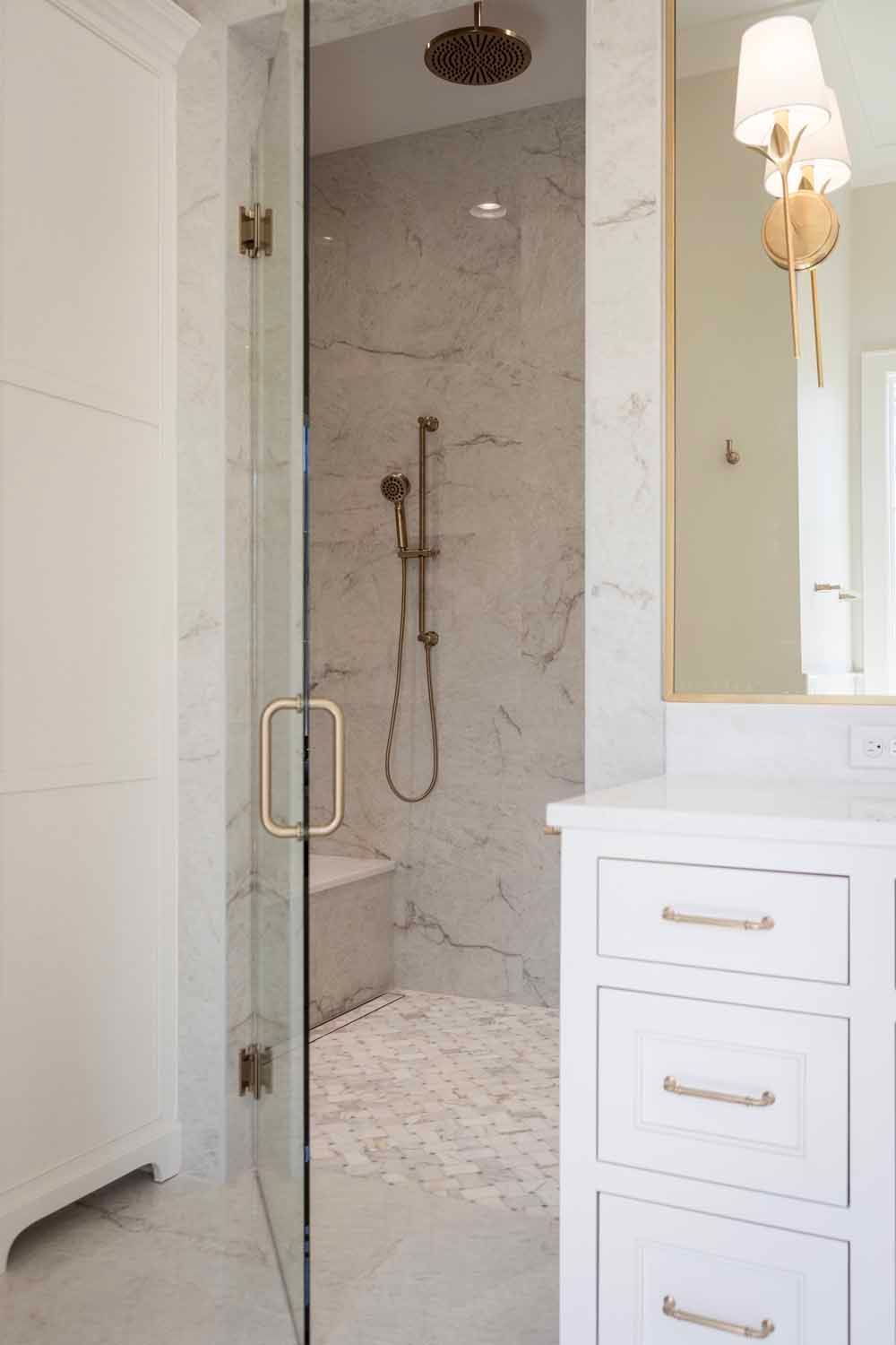A marble-tiled walk-in shower with gold fixtures next to a white vanity with gold drawer pulls and a matching wall sconce.
