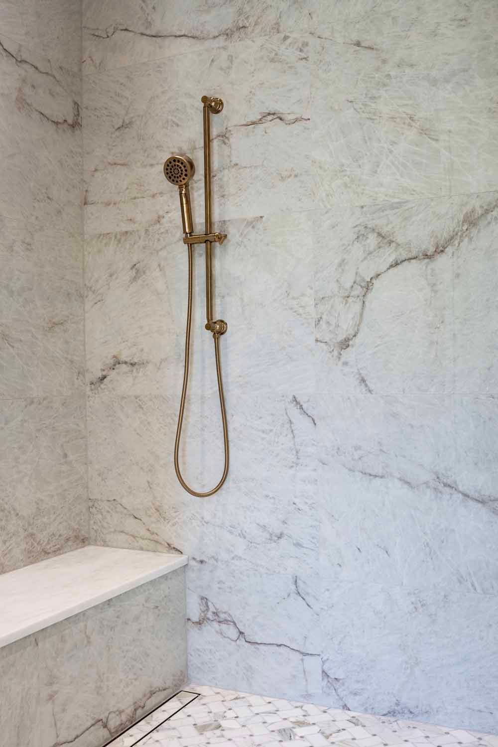 A modern walk-in shower with white marble walls, a matching bench, and a brushed gold handheld shower fixture.