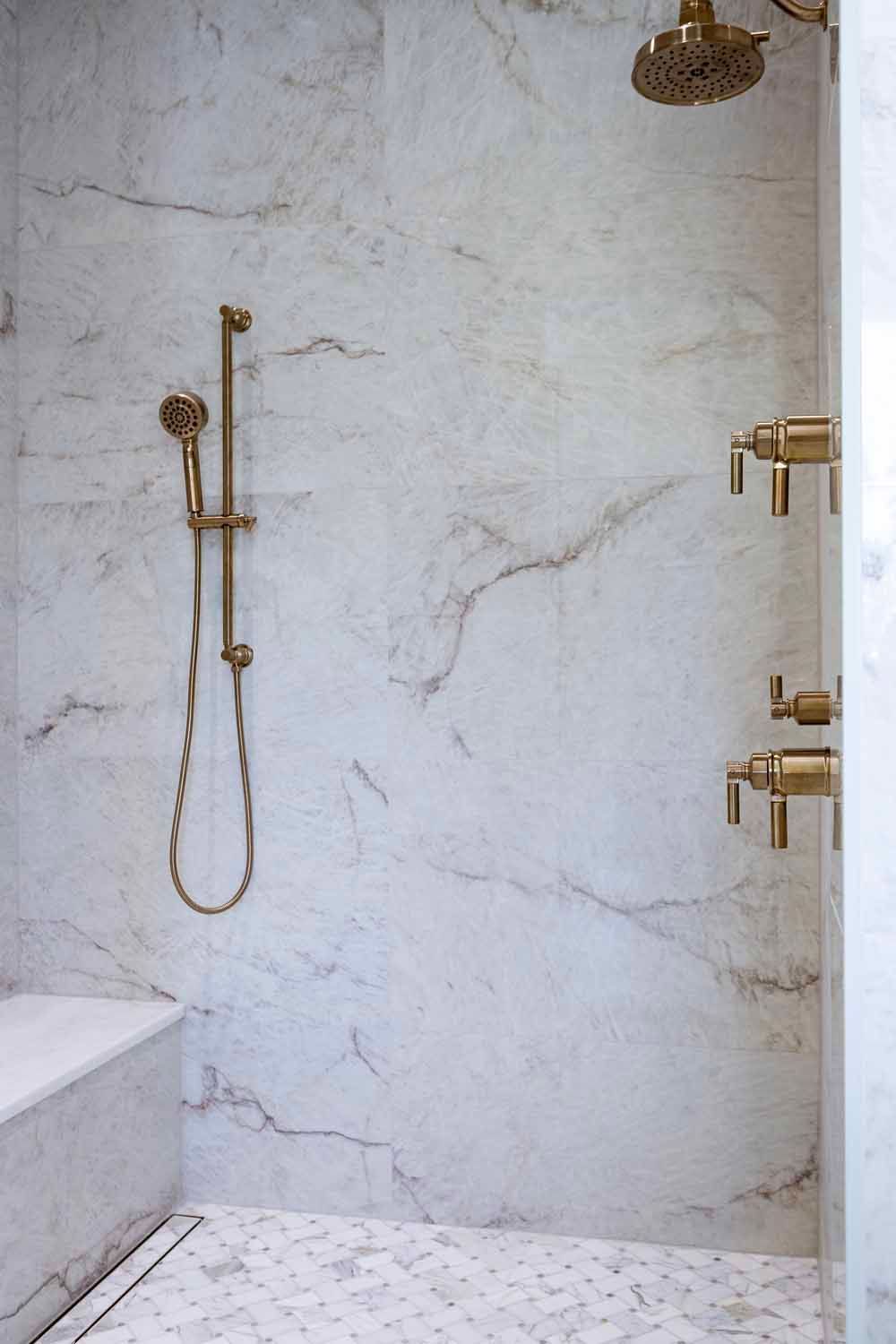 A shower with white marble walls and a tiled floor, featuring gold-toned hardware, including a showerhead and controls.