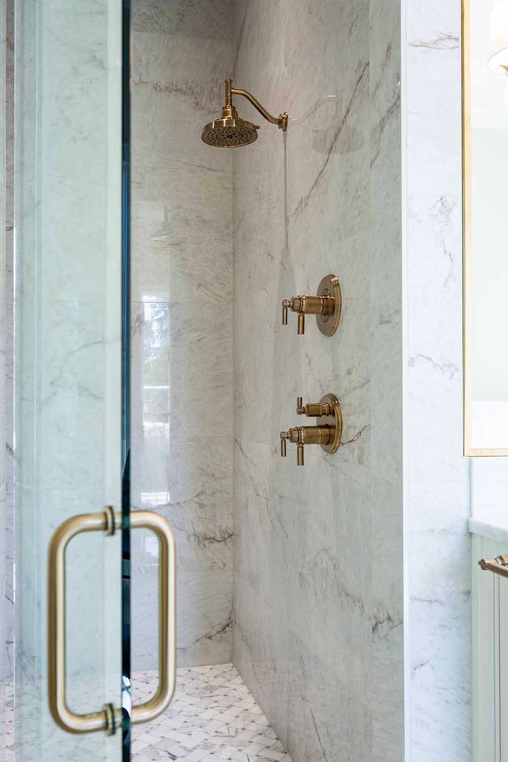 A marble walk-in shower with a glass door, gold hardware, and a brass shower head against white veined tile walls.