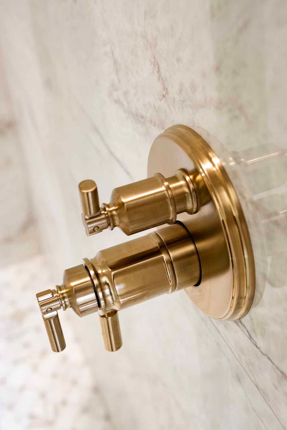 A gold-finished dual-handle shower control valve mounted on a light, textured marble wall.