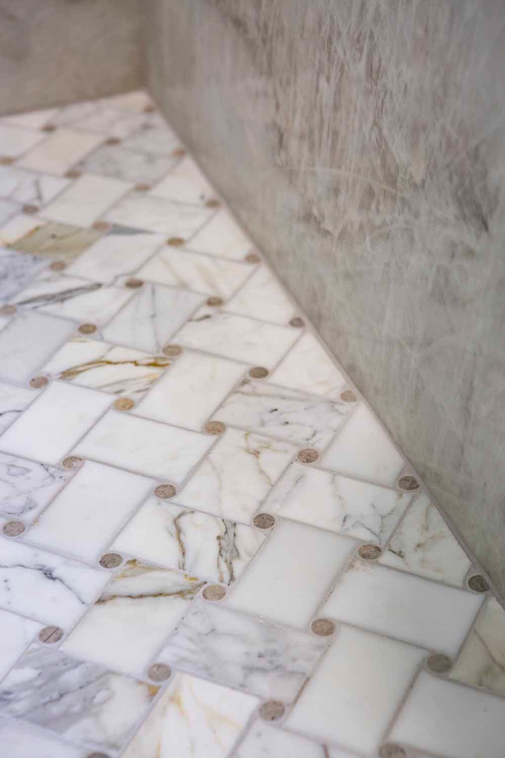 A white marble basketweave tile floor with small circular accents meeting a marble wall.