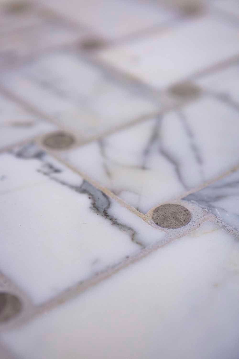 A close-up view of white marble tiles arranged in a grid, accented by small, circular grey stone insets at the corners.