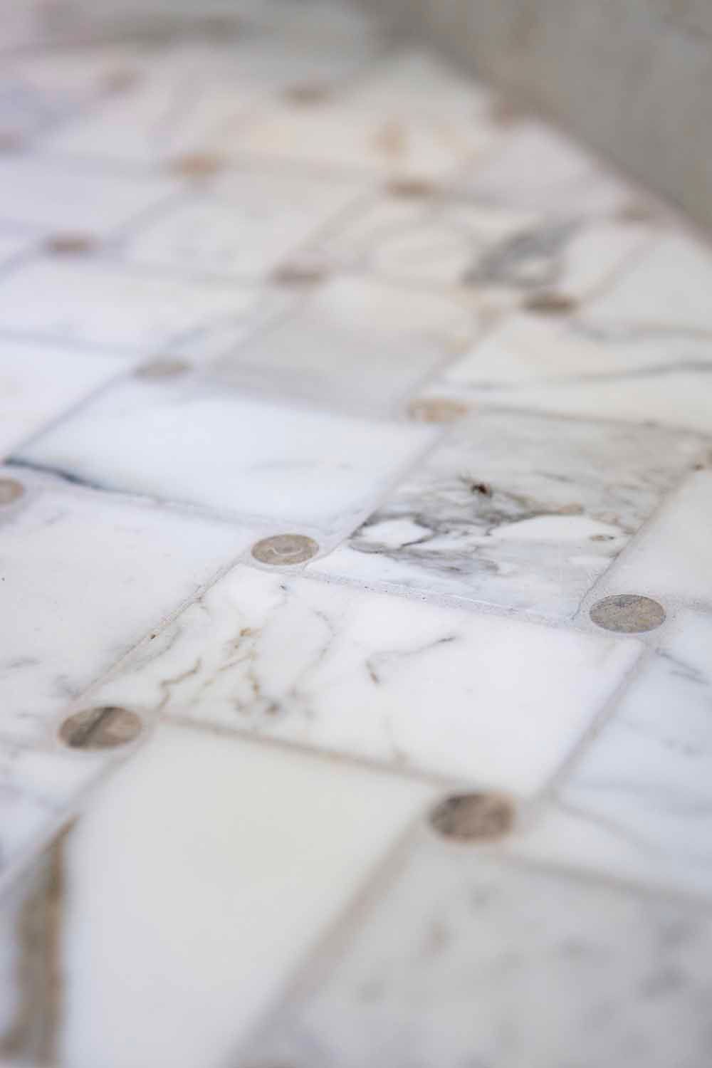 Close-up of white marble floor tiles with grey veins and small, circular, brown stone inlays at the corners.