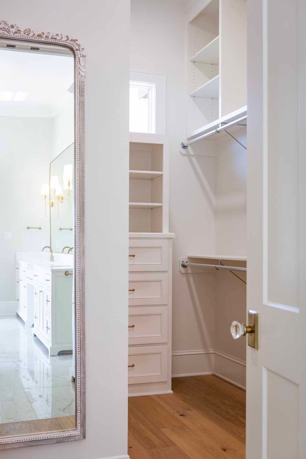 A white walk-in closet with shelves and drawers, viewed through a doorway with a full-length mirror on the adjacent wall.