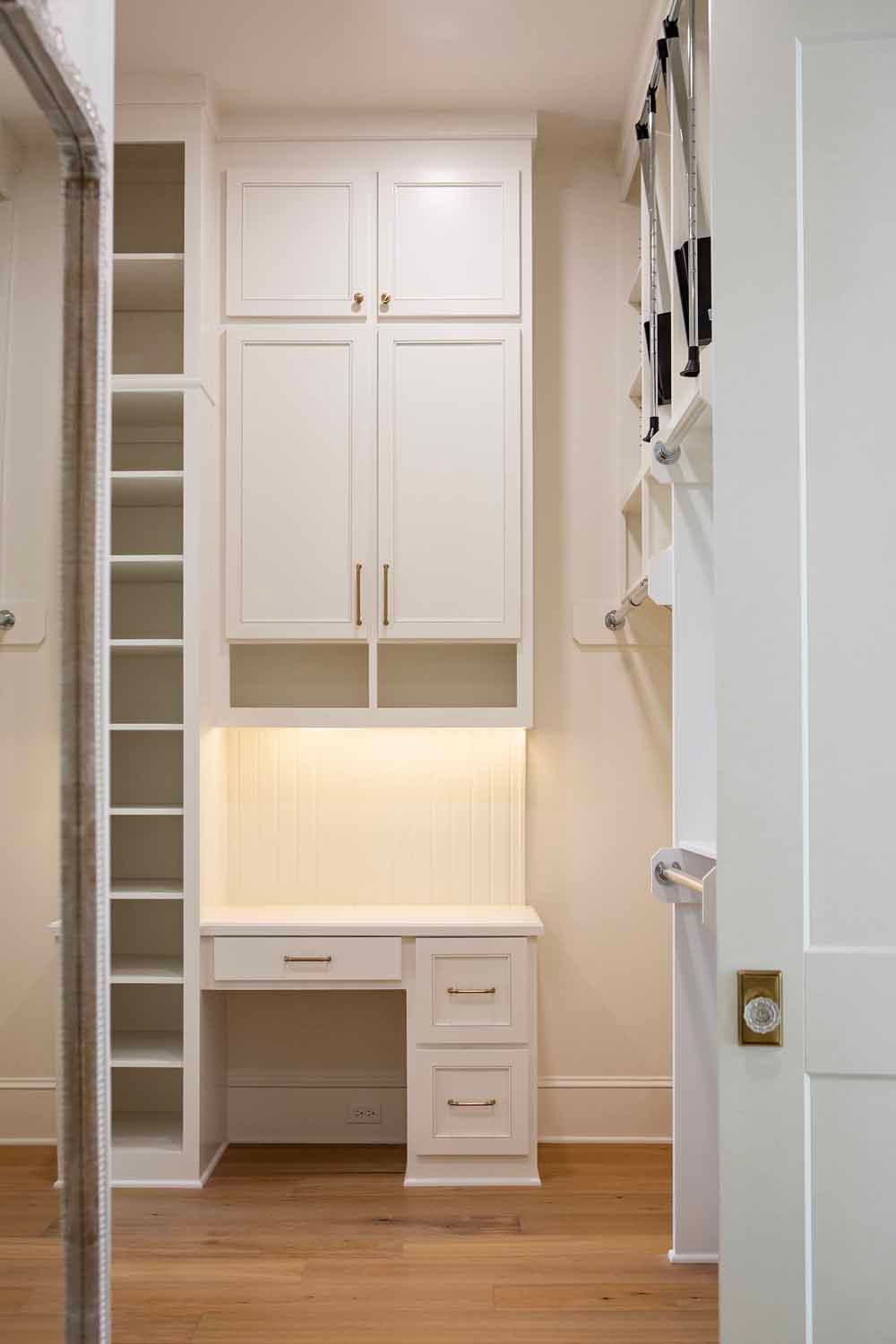 A white built-in desk unit with overhead cabinets, open shelving, and a small drawer set in a neutral-toned room.