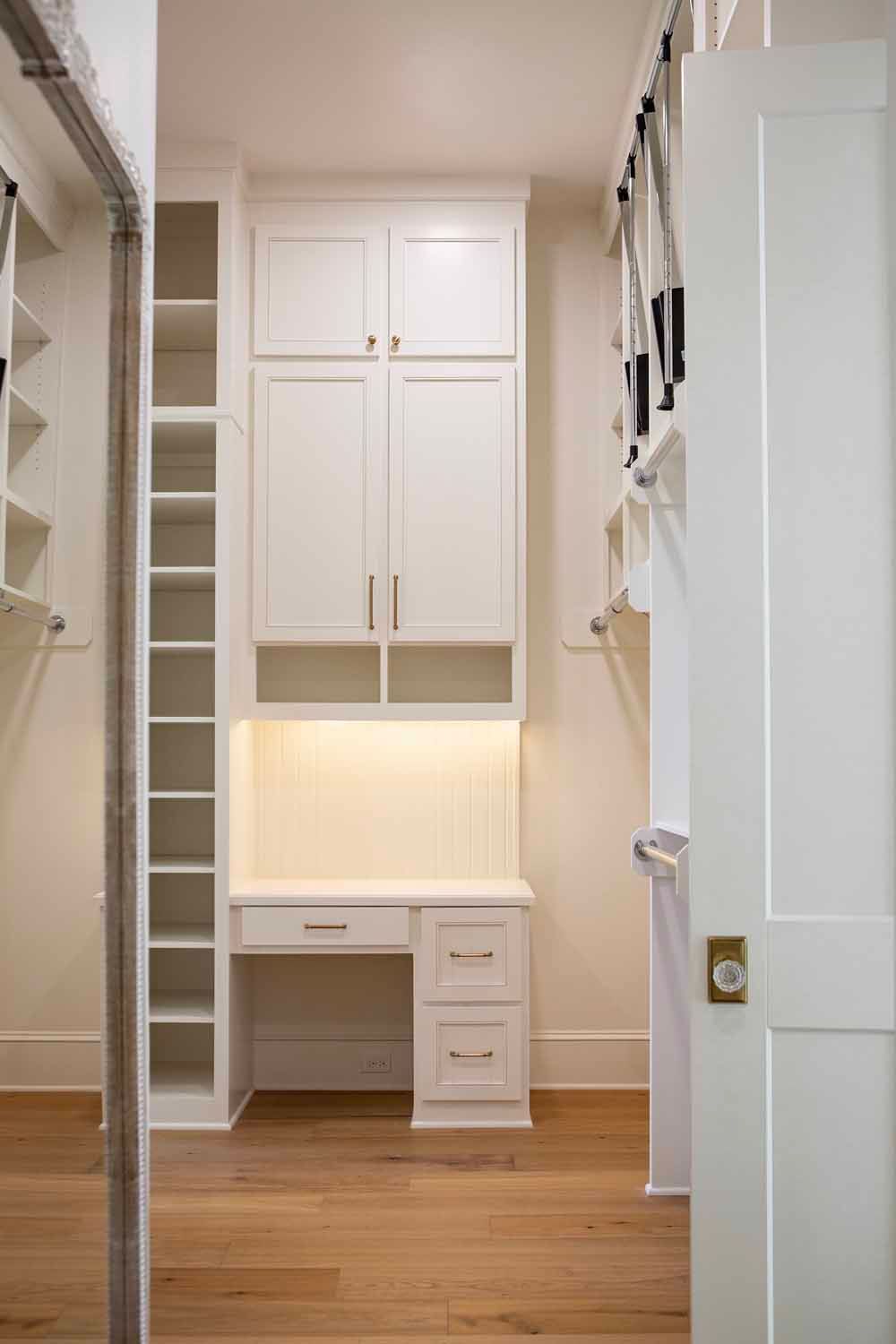 A walk-in closet featuring white cabinetry, a built-in desk with lighting, open shelving, and wood floors.