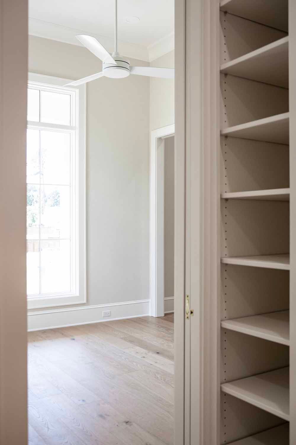 A white ceiling fan hangs above a sunlit room viewed from a closet doorway with stacked empty white shelves.