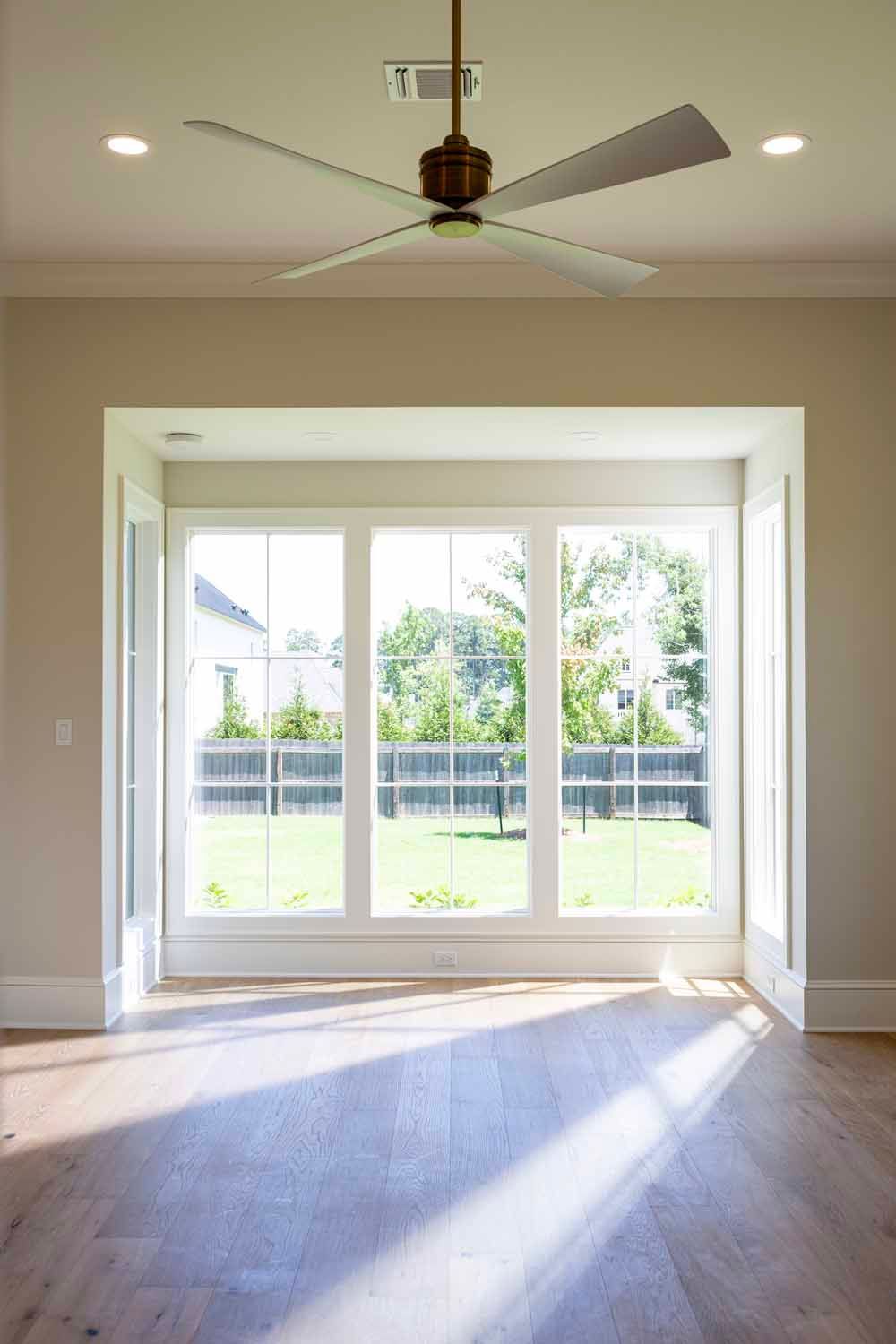 A room with light wood floors, a ceiling fan, and large windows looking out onto a backyard with a fence and trees.
